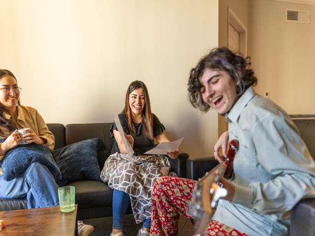 Students sitting on couches in their housing room smiling and one is playing the guitar.