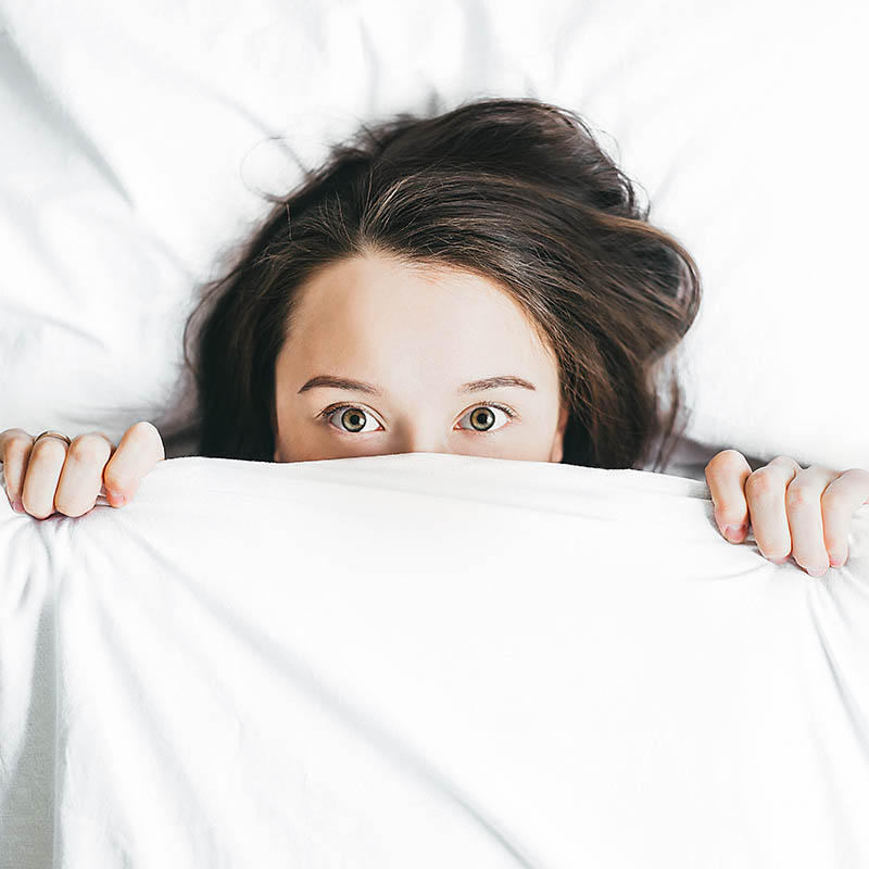Woman laying in white bed with sheets pulled up over her nose. She's making mischievous eyes