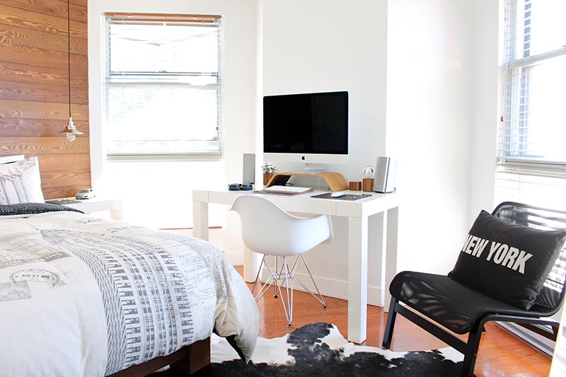 A dorm room with light colors, a desktop computer and a bed with white sheets