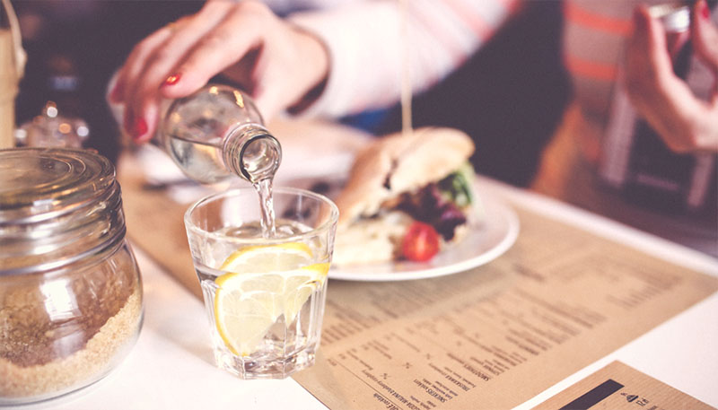 A person serving water with fruit in it from a glass bottle into a glass on a table