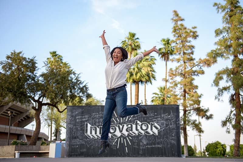 A girl leaping enthusiastically in front of a chalkboard that says "innovation" and trees are behind it